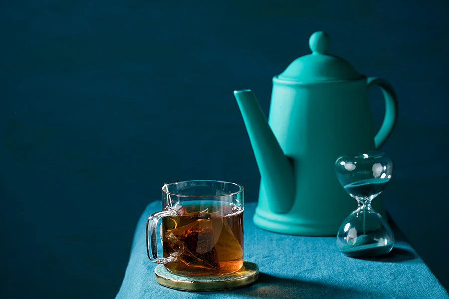 A cup of tea sits next to a blue teapot and an hourglass Two cups of tea sit on a saucer on a table