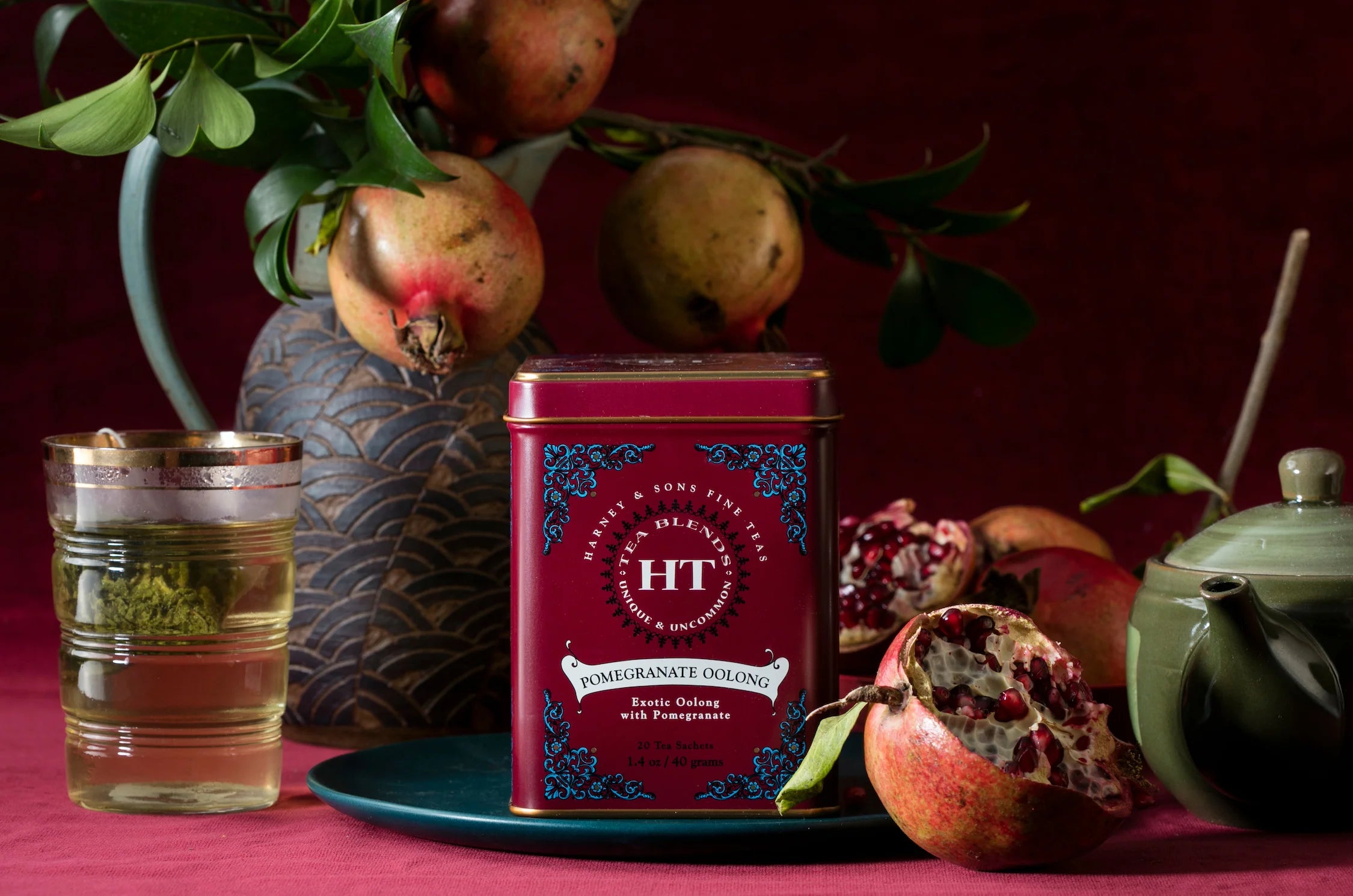 A tin of Harney & Sons Pomegranate Oolong tea, surrounded by pomegranates, a teapot, and a glass of brewed tea.